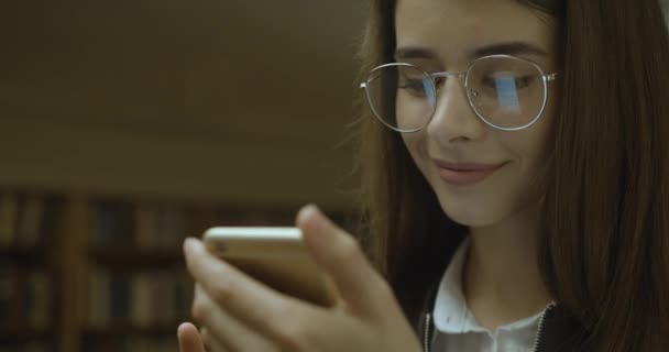 Akıllı telefonla çalışan bir kız öğrenci. Büyüleyici kız kütüphanede dururken telefonunda bir şeyler okuyor. — Stok video