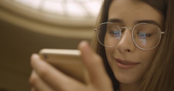 Estudiante femenina, trabajando con smartphone. Chica encantadora lee algo en su teléfono de pie en la biblioteca — Vídeo de stock