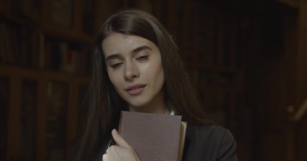 Kvinnlig student, bibliotek. Charmig ung kvinna tittar rakt in i kameran och ler stående i det mörka rummet och kramar sin bok — Stockvideo