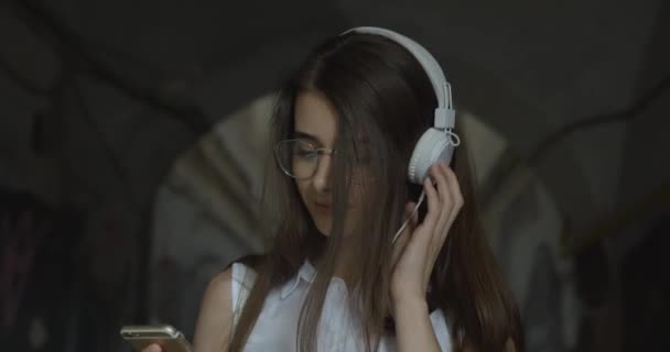 Encantadora chica de gafas pasea por la ciudad y escucha la música en sus auriculares — Vídeos de Stock