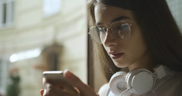 Chica joven en gafas lee algo en su teléfono inteligente — Vídeos de Stock