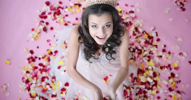 스튜디오에서 모델 포즈하는 거요. 머리에 곱슬곱슬 한 털 과 왕관을 쓰고 바닥에 앉아 있는 장미꽃 꽃잎 위로 올라가는 모델이다. 긍정적 인 감정 — 비디오