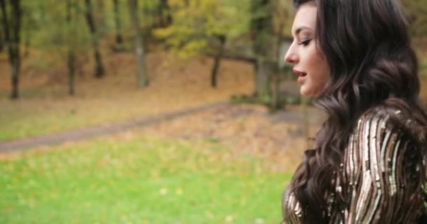 4k. Frau in der Stadt. Attraktives Model im glitzernden Abendkleid spaziert im Herbstpark über den Pass und schüttelt ihre langen brünetten Haare — Stockvideo