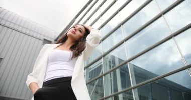 4 bin. Yavaş ve hızlı çekim. Süslü giyinmiş, siyah beyaz elbiseli genç bir iş kadını dışarıda modern cam bir binanın önünde poz veriyor.