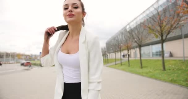 4k. Movimiento lento y rápido. Elegante joven mujer de negocios vestida con ropa blanca y negra camina a lo largo del paso ante un moderno edificio de vidrio, se da la vuelta y sacude su cabello — Vídeos de Stock