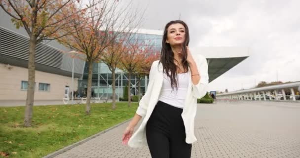 4k. Movimento lento e rápido. Fantasia vestida jovem mulher de negócios em roupas pretas e brancas caminha ao longo do passo antes de um edifício de vidro moderno, vira-se, balança o cabelo e olha diretamente para o — Vídeo de Stock