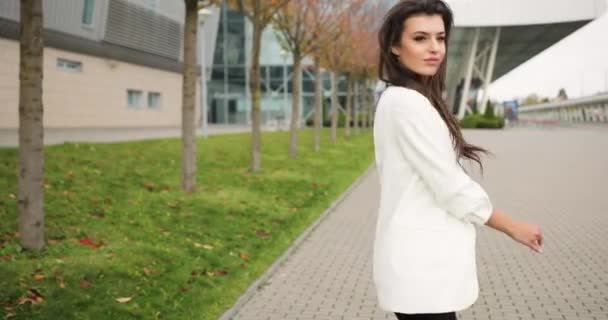 4k. Movimiento lento y rápido. Mujer de negocios joven vestida elegante en ropa blanca y negra camina a lo largo del paso antes de un edificio de vidrio moderno, se da la vuelta, sacude su cabello y mira directamente a la — Vídeos de Stock