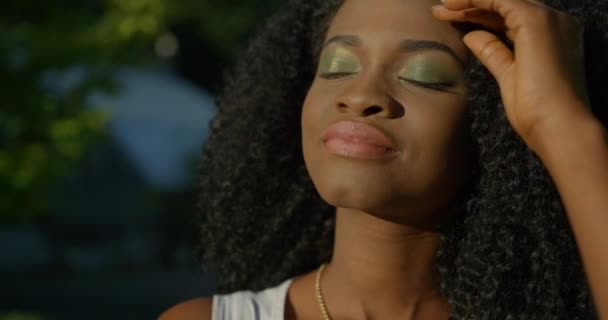 Yeşil göz farı ve tarafsız ruj sürmüş barışçıl çekici Afro-Amerikalı kız hafifçe gülümsüyor ve siyah kıvırcık saçlarını düzeltiyor. Yakın plan portre, 4k. — Stok video