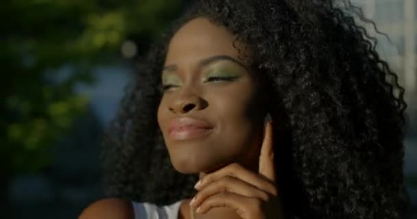 Ritratto dell'adorabile giovane afro-americana con ombretti verdi, rossetto neutro e vista tranquilla. Sta correggendo i suoi capelli neri ricci, scuotendo la testa e sorridendo. — Video Stock