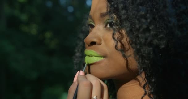 La vista lateral de cerca del maquillador profesional aplicando el lápiz labial verde en la encantadora chica afro-americana con sombras de ojos verdes y cabello rizado en el fondo verde — Vídeos de Stock