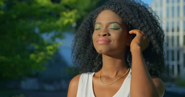 Retrato de cerca de la pacífica y hermosa afro-americana con sombras de ojos verdes y lápiz labial neutro corrigiendo su cabello rizado mientras disfruta del clima con los ojos cerrados — Vídeos de Stock