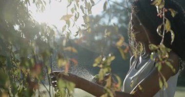 Doğal makyajlı gülümseyen Afro-Amerikalı kız güneş kremlerinin arka planında yeşil plastik tetik spreyi kullanarak açık hava bitkileriyle ilgileniyor. 4k, ağır çekim..