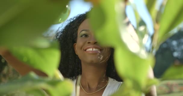 Primer plano retrato a través de las plantas. Encantadora mujer afroamericana con lápiz labial neutro y sombras de ojos verdes sonríe pacíficamente mientras usa el spray de plástico verde. — Vídeo de stock