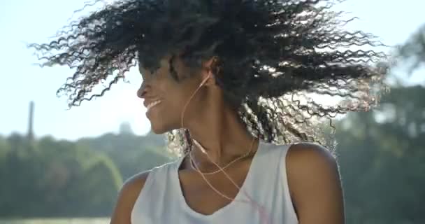 Ritratto della bella ragazza afro-americana con ombretti verdi che sorridono, scuotono i capelli ricci e ballano ascoltando musica con le cuffie sullo sfondo del sole. — Video Stock