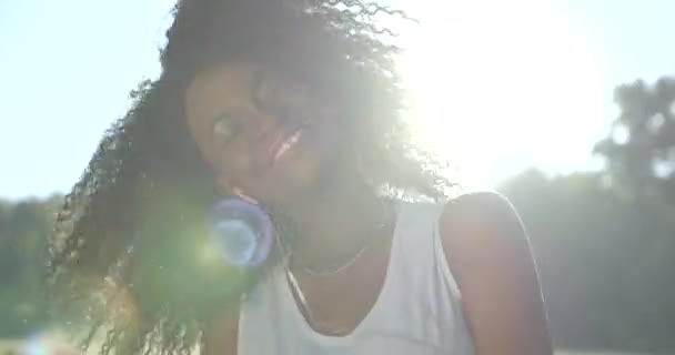 Енергетична афро-американська дівчина з нейтральною помадою і зеленими тінями очей посміхається і активно трясе її темне кучеряве волосся через музику в її навушниках на фоні сонячних променів . — стокове відео