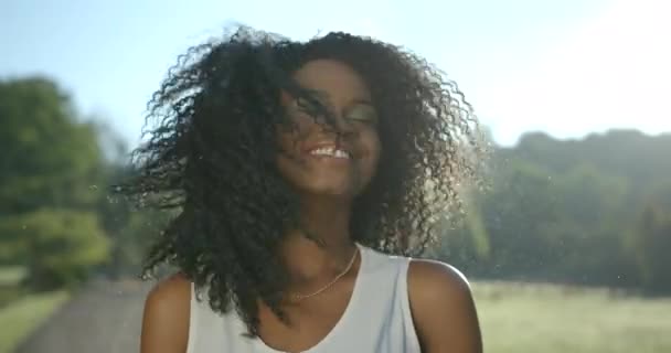 Divertida chica afroamericana enérgica con sombras de ojos verdes está sonriendo, temblando alegremente y tocando emocionalmente su cabello rizado. Retrato exterior. — Vídeo de stock