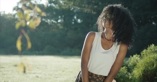 Menina afro-americana adorável com sorriso encantador e sombras de olhos verdes está tremendo seu cabelo encaracolado e mãos enquanto dança energeticamente por causa da música nos fones de ouvido. Retrato exterior. — Vídeo de Stock