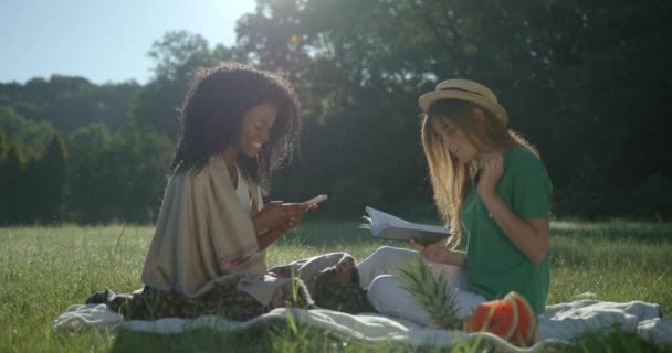 Dos hermosas novias de carreras múltiples están haciendo el picnic en el parque. El afro-americano está chateando a través del teléfono móvil mientras que el caucásico está comiendo las uvas y leyendo el libro. — Vídeos de Stock
