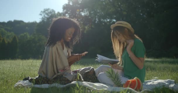 Két gyönyörű, többnemzetiségű barátnő eltöltésére egy pikniken. A mosolygó kaukázusi elolvassa a könyvet és megeszi a szőlőt, miközben az afrikai lány SMS-ezik és böngészi a mobiltelefonját.. — Stock videók