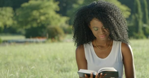 Вид спереди молодой африканской девушки с очаровательной улыбкой и тенями зеленых глаз, читающей книгу на лужайке. — стоковое видео