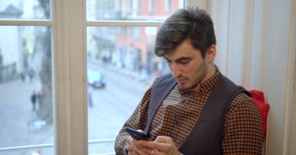 Крупным планом портрет красивого серьезного мужчины, который пишет смс, болтает, гуляет по сети с помощью мобильного телефона, сидя на подоконнике. — стоковое видео