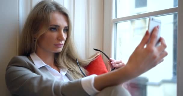 Close-up portret van de mooie blonde zakenvrouw met bril selfies nemen met verschillende emoties terwijl zitten op de vensterbank. — Stockvideo