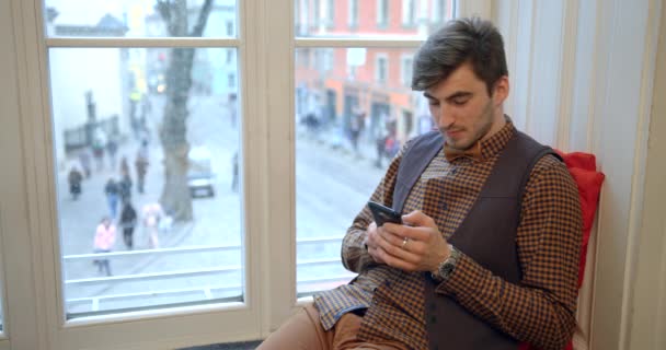 Сосредоточенный молодой привлекательный мужчина чатится, пишет смс и просматривает сеть через мобильный телефон, сидя на подоконнике. 4k. — стоковое видео