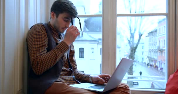 A fáradt üzletember leveszi a szemüvegét, megdörzsöli a szemét, és tovább dolgozik a laptopon, miközben az ablakpárkányon ül. Aztán kinézett az ablakon.. — Stock videók