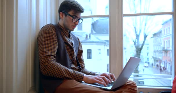 Oldalnézet a fiatal üzletember szemüveges összpontosít a munka a laptop, miközben ül az ablakpárkányon, akkor néz ki az ablakon keresztül. — Stock videók