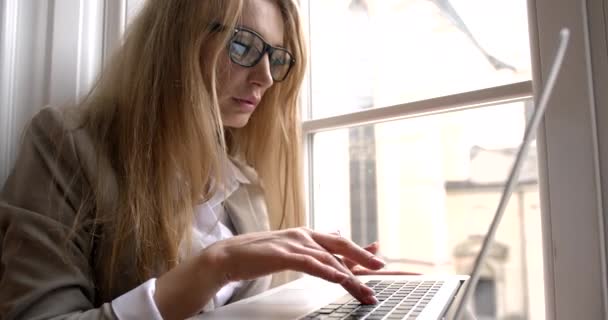 Närbild sida porträtt av den allvarliga blonda affärskvinna bär glasögon och arbetar på den bärbara datorn medan du sitter på fönsterbrädan. Reflektion av grafik i glasögonen. — Stockvideo