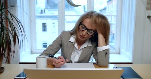 Vista frontal de la mujer de negocios rubia sobrecargada de trabajo escribiendo algo, tomando té y quitándose las gafas en el lugar de trabajo. — Vídeos de Stock