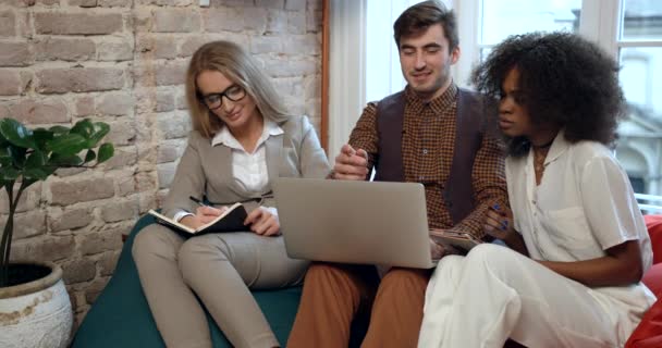 Team work of three multirace businesspeople working on the laptop and laughing. The young blonde woman is writing something down. — Stock Video