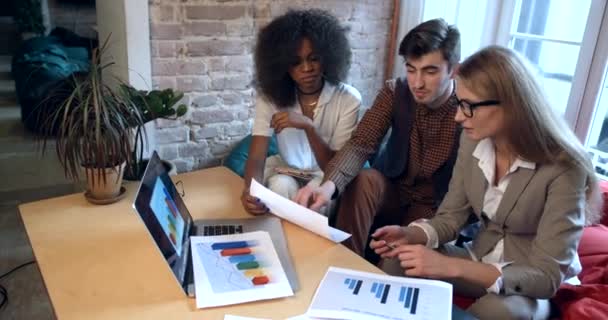 Discusión activa de tres colegas multirracistas que trabajan en el portátil y diagramas de negocios y gráficos en el lugar de trabajo. Trabajo en equipo. Concepto de oficina. — Vídeo de stock