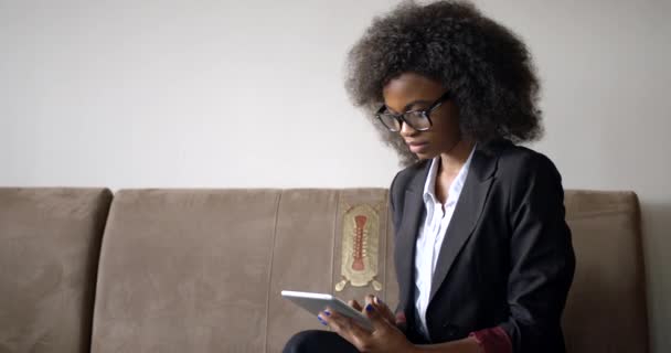 魅力的な幸せなアフロアメリカのビジネスマンの女性は、ソファに座っている間、タブレットを介してインターネットを笑顔とチャット、テキストメッセージ、サーフィンしています。4k. — ストック動画