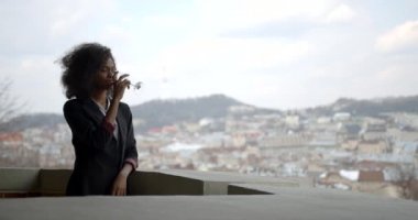 Çekici Afro-Amerikalı takım elbiseli kadın balkonda dikilirken şarabın ve manzaranın tadını çıkarıyor..
