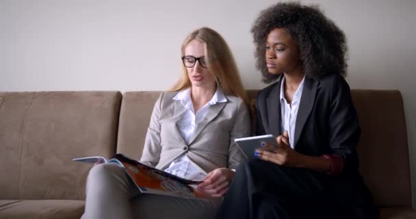 Encantadora mujer de negocios rubia con gafas está mostrando la revista a la hermosa trabajadora afroamericana con la tableta mientras está sentada en el sofá. — Vídeos de Stock