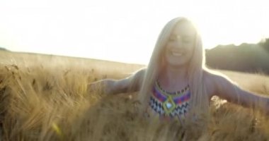Doğal makyajlı neşeli, gülümseyen sarışın kız altın tarlada vakit geçirmenin tadını çıkarıyor. Mutlu bir şekilde dönüyor ve güneşteki altın buğdaylara dokunuyor..