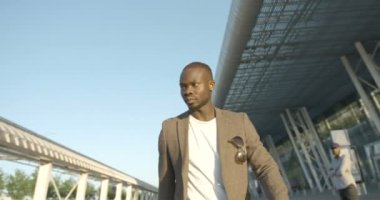 Kendine güveni tam modaya uygun Afrikalı bir adamın, güneşli caddede yürüdüğünü gösteren bir kamera. 4k görüntü.