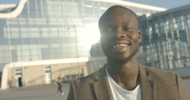 Emotioneel close-up portret van de charmante Afrikaanse man met een mooie witte glimlach lachend over glazen bouwachtergrond tijdens de zonnige dag. 4k-beelden. — Stockvideo