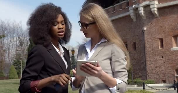 Zakelijk gesprek tussen twee jonge multiculturele zakenvrouwen in pakken die de tablet gebruiken. Close-up outdoor portret — Stockvideo