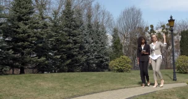 Vista completa de dos mujeres de negocios mixesd raza en trajes discutiendo los problemas de negocios mientras camina por la calle. El afroamericano sostiene la tableta. — Vídeo de stock