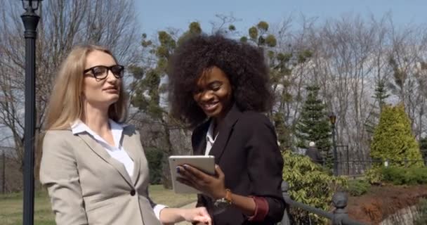 Twee vrolijke gemengde ras zakenvrouwen zijn actief praten en lachen tijdens het surfen op het net en het bekijken van video via de tablet. Close-up outdoor portret. — Stockvideo