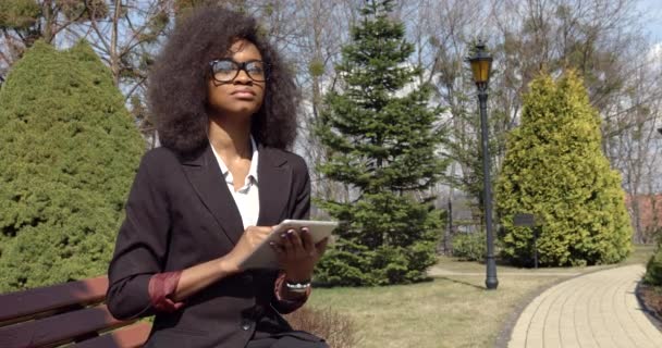 这位年轻美丽的非洲裔美国女商人正在摘下眼镜，一边坐在长椅上一边通过平板电脑聊天，一边欣赏风景. — 图库视频影像