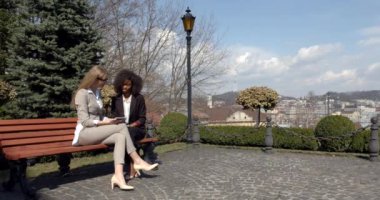 İki çok ırklı genç iş kadını, manzara manzarasının arka planındaki bankta otururken tableti konuşuyor ve kullanıyor..