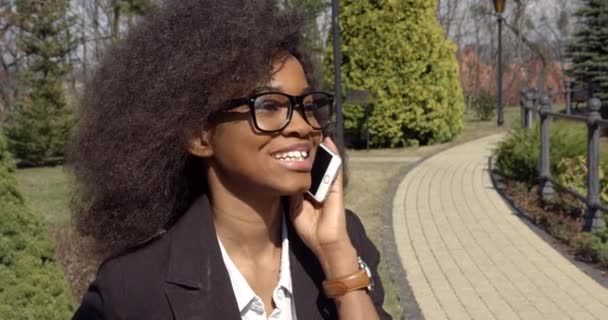 Side portrait of the young attractive afro-american business woman with dark curly hair wearing glasses lachend while talking via the mobile phone in the park. — Stockvideo