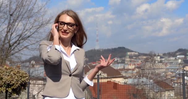 Hermosa mujer de negocios rubia feliz en gafas está sonriendo mientras habla a través del teléfono móvil en el fondo del panorama de la ciudad. — Vídeos de Stock