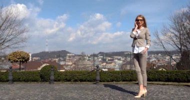 Gözlüklü ve bej takım elbiseli çekici sarışın iş kadını şehir manzarasının arka planında cep telefonuyla neşeli bir şekilde konuşuyor..