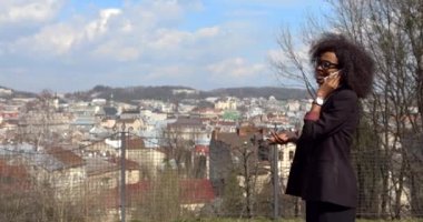 Gözlüklü, siyah takım elbiseli, gülümseyen ve cep telefonuyla konuşurken hareket eden Afrikalı güzel iş kadınının yarım boy portresi. Açık hava görünümü.