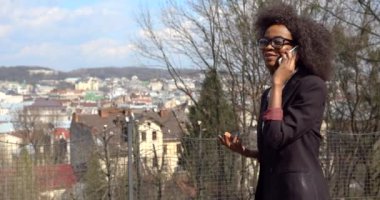 Takım elbiseli ve gözlüklü güzel Afrikalı iş kadını cep telefonuyla konuşurken sokakta gülüp dolaşıyor. Yarım boy portre. 4k.