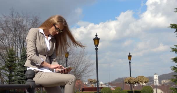 Urocza blond bizneswoman w okularach rozmawia, pisze i przegląda przez tablet, a następnie odwraca wzrok i używa gadżetu siedząc na żelaznej barierze. — Wideo stockowe